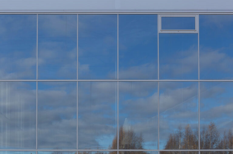 Reflektion eines blauen Himmels mit Wolken und Bäumen auf einer großen Glasfassade.